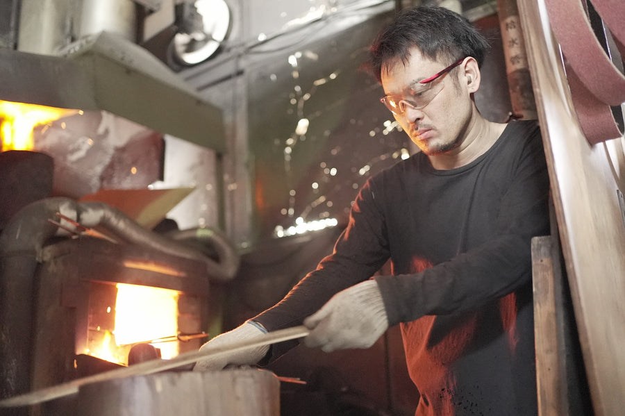 Satoshi Nakagawa at the grinding wheel, forge glowing behind.